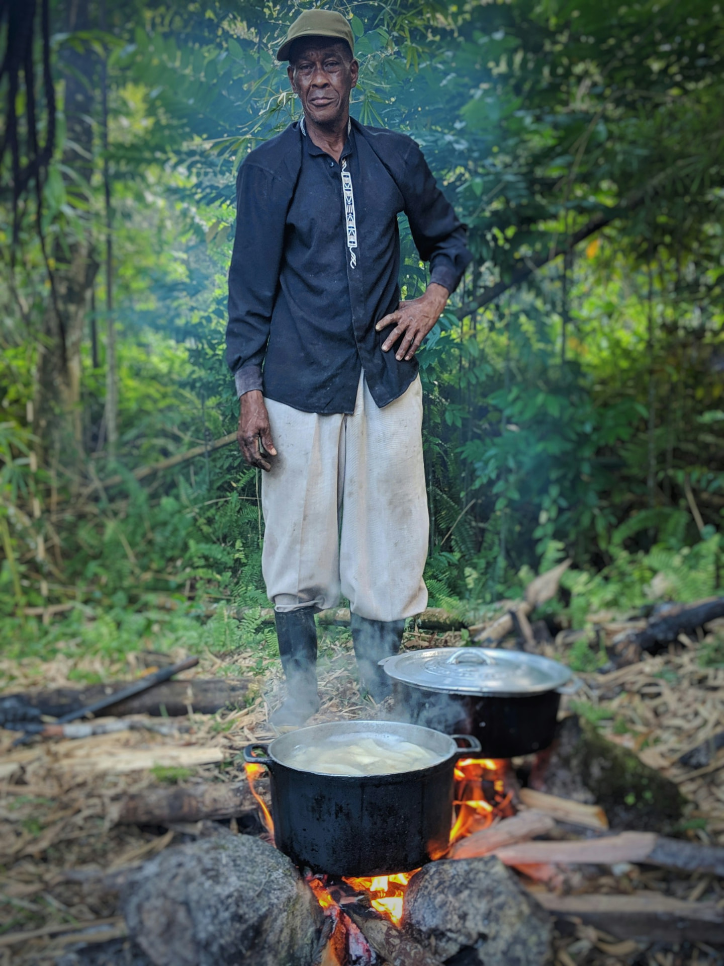 A cook stands over two dutch pots on an open wood fire in the Jamaican bush